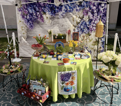 A vibrant and elegantly decorated Nowruz Haft-Sin table set up under a white tent at the Beverly Hills Farmers Market. The table features a lime green cloth, floral backdrops, and traditional Persian symbolic items like apples, sprouts, and goldfish, representing renewal and luxury living in Beverly Hills.