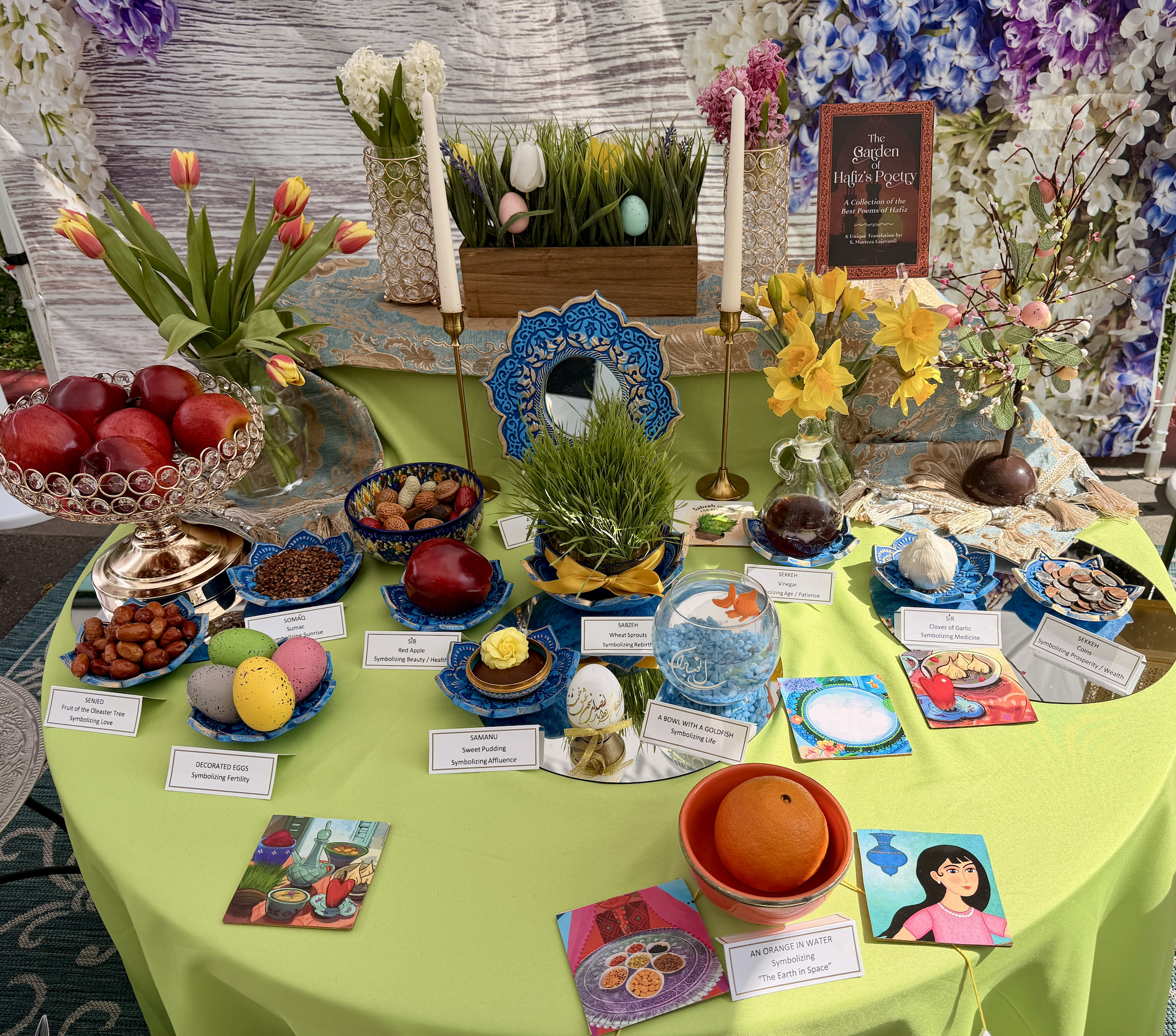 Close-up of a traditional Persian Haft-Sin arrangement in Beverly Hills, showing symbolic "S" items: Sabzeh (sprouts), Sib (apples), Serkeh (vinegar), and Samanu (pudding) in ornate blue bowls. A book of Hafiz poetry and a blue mosaic mirror reflect the sophisticated cultural heritage of the 90210 community.