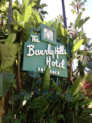 Iconic green and white entrance sign of The Beverly Hills Hotel surrounded by lush tropical banana leaves and palm trees on Sunset Boulevard.