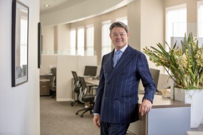 Headshot of Christophe Choo, a top real estate agent and licensed broker since 1989, in a tailored navy pinstripe suit, leaning confidently in a Beverly Hills luxury office setting with large window views