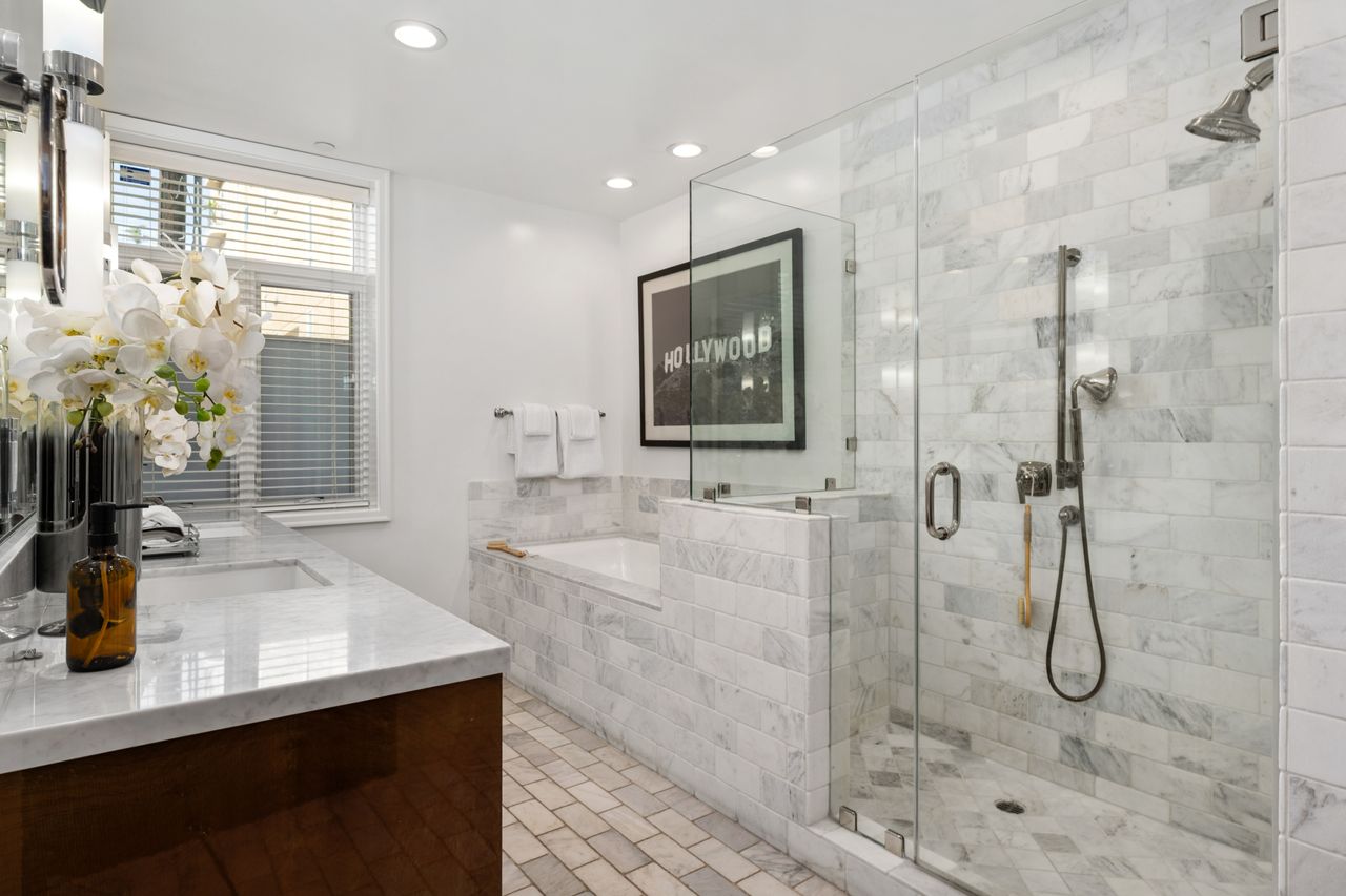 Spa-inspired primary bathroom featuring a seamless glass shower stall, separate soaking tub, marble brick tile walls, and a marble vanity countertop with modern dual sinks.