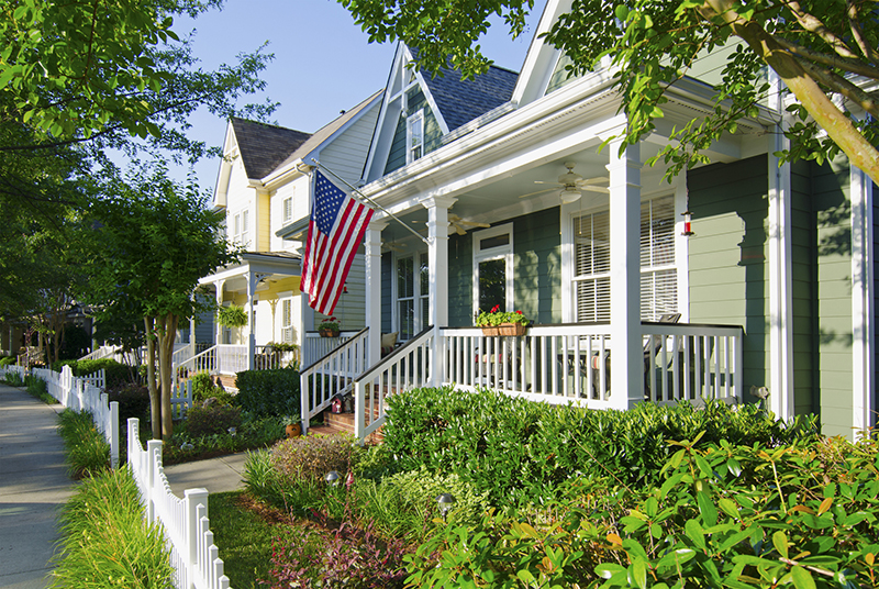 Fort Mill, South Carolina, USA - June 14, 2014: The American Dream is pictured in this iconic image of a row of new, Victorian-style homes with a white picket fence in the Baxter Village neighborhood development located south of Charlotte, North Carolina. Most American homes are now built on smaller lots with sidewalks and tree-lined streets in the suburbs of large cities. An American flag hangs from the front porch in celebration to honor an upcoming holiday. Many Americans express patriotism by flying Old Glory, not only on holidays, but all year long.