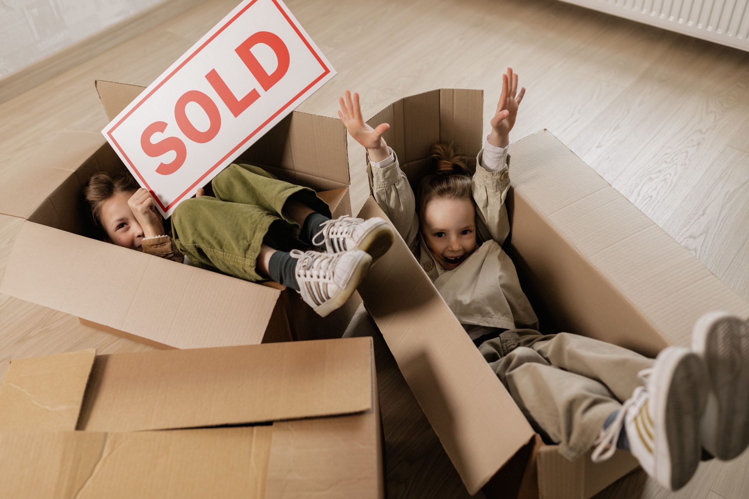 Two children celebrating in moving boxes while holding a SOLD sign, representing the successful completion of a home sale and purchase.