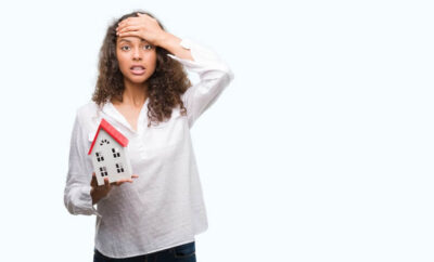 a woman holding a mini model house while other hand on her forehead