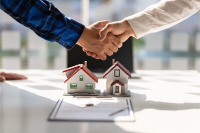 Two people shake hands over a desk with miniature house models and a document, symbolizing a real estate transaction or agreement.