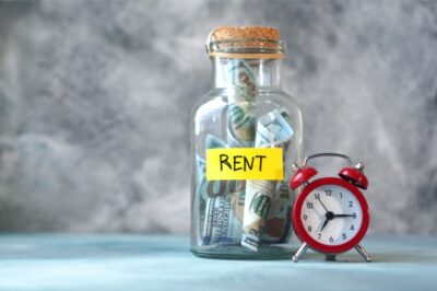 A glass jar labeled "RENT" filled with rolled dollar bills sits next to a small red alarm clock,