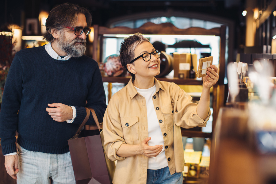 Mature couple choosing products at a boutique shop. Woman smiling while looking at an item, man holding a shopping bag