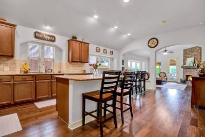 Open floor plan kitchen and living area with granite island, bar seating, wood floors, and brick fireplace in a San Marcos TX home.