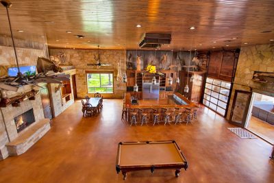 Texas Hill Country estate San Marcos TX entertainment hall with indoor bar, pool table, fireplace, interior guest quarters, and access to outdoor swimming pool, outdoor bar, and large stone BBQ pit