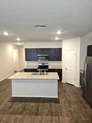 Open kitchen with large granite island, dark cabinetry, stainless steel appliances, and wood-look flooring in newer Seguin home