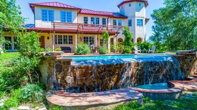 Infinity-edge pool with stone waterfall feature overlooking a custom estate and landscaped outdoor living area