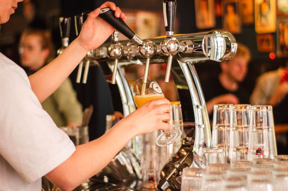 A bartender pours a drink from a tap into a glass, with blurred patrons and stacked glasses in the background.