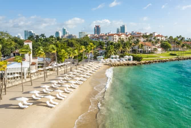 A scenic beach view with white umbrellas, palm trees, and turquoise water, set against a backdrop of city buildings.