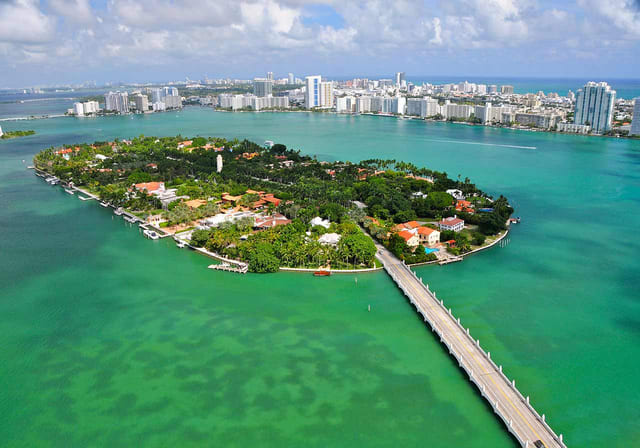 Aerial view of a lush green island surrounded by turquoise waters, with a bridge connecting to the mainland and city skyline in the background.