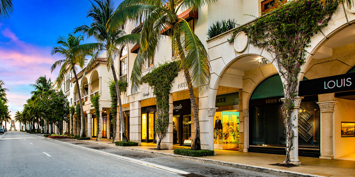 A palm-lined street featuring upscale shops with elegant architecture and a colorful sky at dusk.