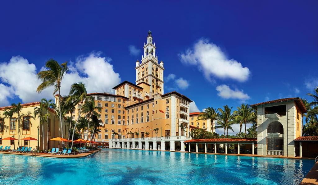 A luxurious hotel with a tall tower, surrounded by palm trees and a large swimming pool under a clear blue sky.