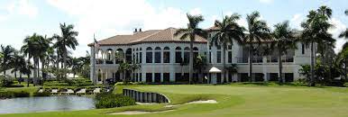 A large, elegant building surrounded by palm trees, overlooking a golf course and a small pond. Bright blue sky in the background.