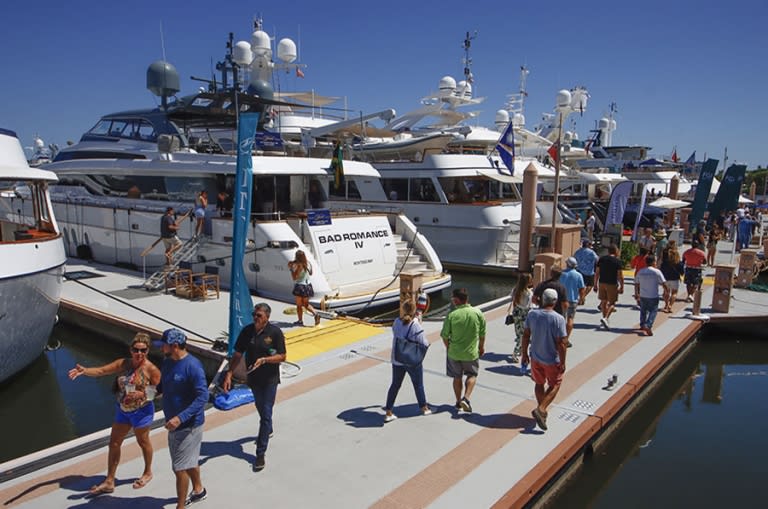A busy marina scene with people walking along docks, surrounded by various yachts and boats under a clear blue sky.