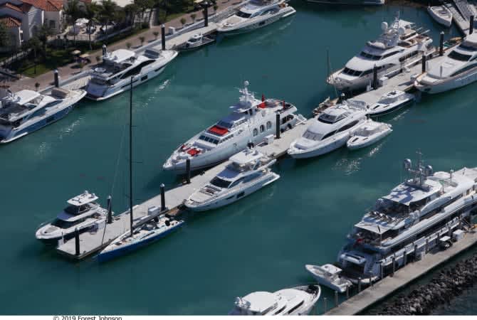 Three luxury yachts are anchored in deep blue water, with one larger yacht prominently in the foreground.