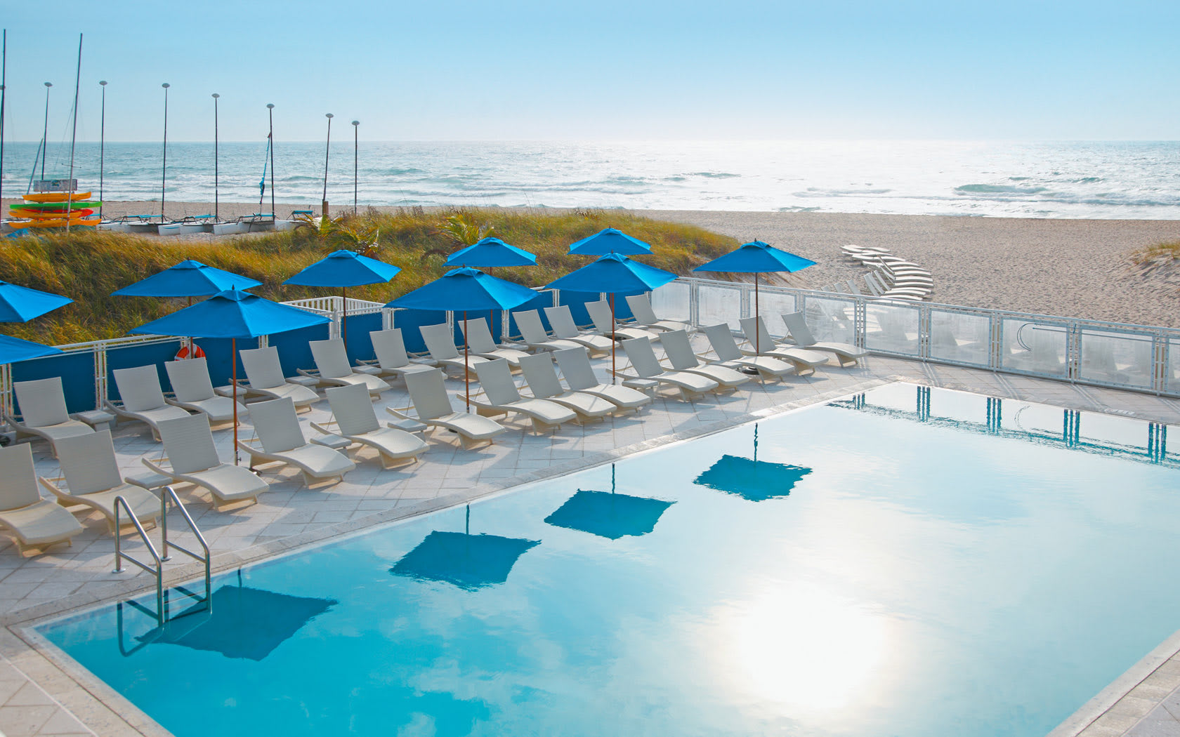 A serene pool area with blue umbrellas and lounge chairs, overlooking a sandy beach and the ocean.
