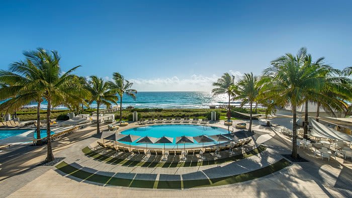 A serene beachfront resort with a circular pool surrounded by palm trees and lounge chairs, overlooking the ocean.