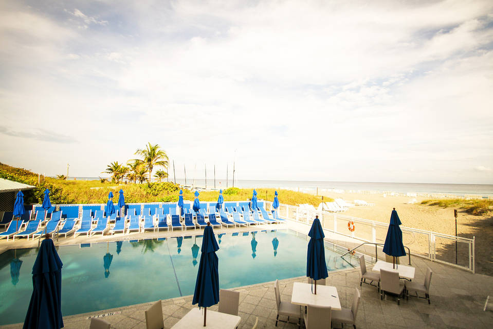 A serene pool area with blue umbrellas and lounge chairs, overlooking a sandy beach and ocean under a partly cloudy sky.