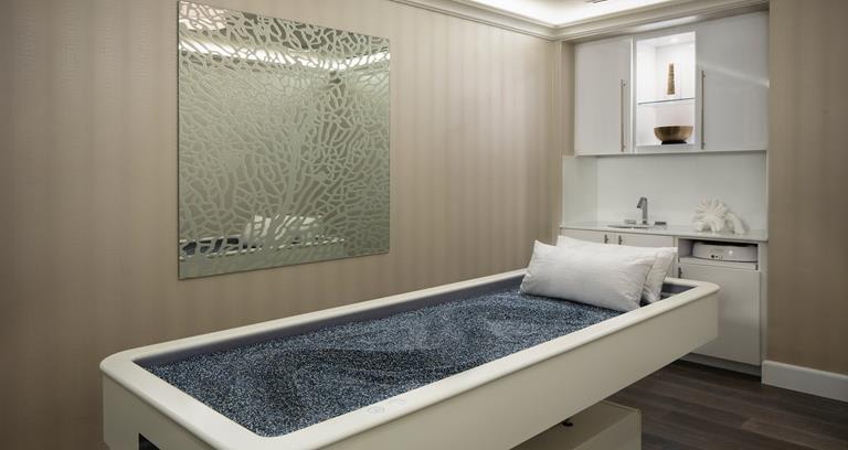 A serene spa room featuring a unique treatment bed filled with dark stones, a decorative mirror, and minimalist cabinetry.