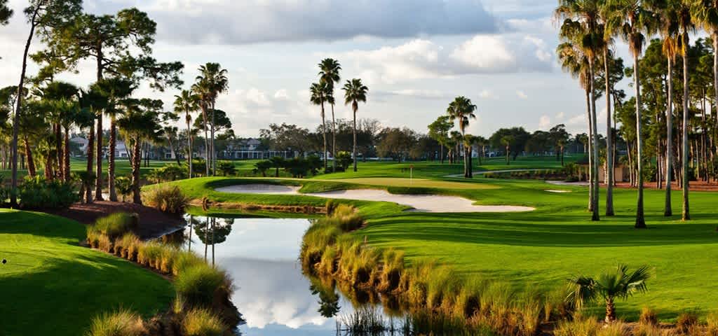 A scenic golf course featuring lush green grass, palm trees, sand traps, and a reflective water hazard under a partly cloudy sky.