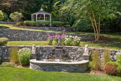 Stone fountain with cascading water in a terraced backyard garden in Lexington MA, featuring stone retaining walls, landscaped greenery, and a white gazebo at the top