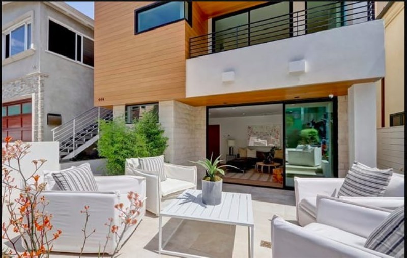 A modern outdoor patio with white furniture, a small table, and greenery, adjacent to a stylish home with large glass doors.