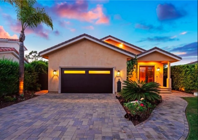 A modern house with a paved driveway, garage, and lush greenery, set against a colorful sunset sky.