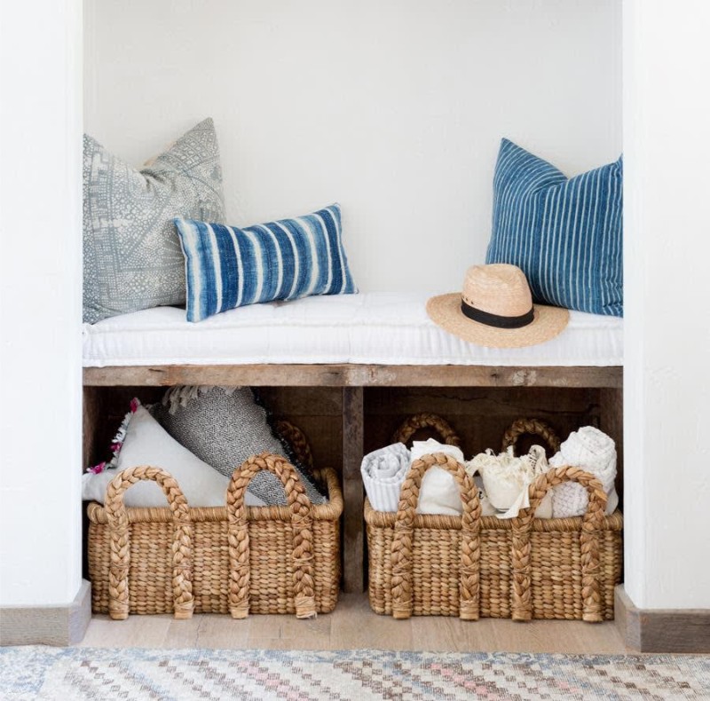 A cozy nook with a white bench, decorative pillows, and woven baskets filled with blankets and towels. A straw hat rests on the bench.