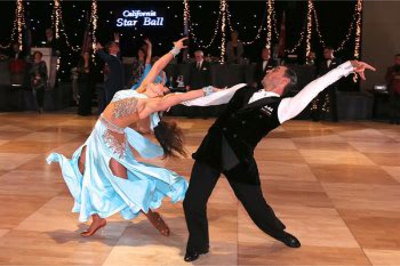 A couple performs a dynamic dance on a wooden floor, showcasing elegant movements in formal attire. Lights and decorations are in the background.