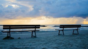 Image is of two benches on the beach facing the ocean. Image is of two benches on the beach facing the ocean.
