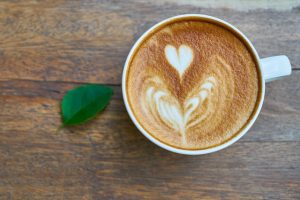 Image is of a cup of coffee with a green leave next to it on a wooden table.