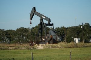 Image is an oil derrick in a field with trees behind it.