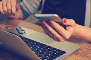 Image is a a woman at a desk with a lap top looking at her iPhone.