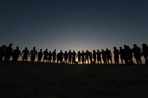 Image is a line of people holding hands, backlit and silhouetted by the setting sun. Image is a line of people holding hands, backlit and silhouetted by the setting sun.