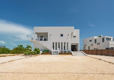 Modern two-story white waterfront estate with exterior staircase, custom architecture, and wide gravel driveway in Tarpon Bay, Ambergris Caye.