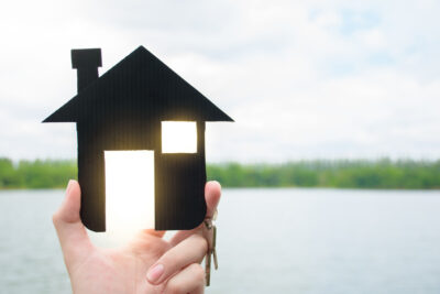 Man hand holding miniature black house model building with keys and soft light on lake and green forest background over sky for concept of home insurance and real estate.