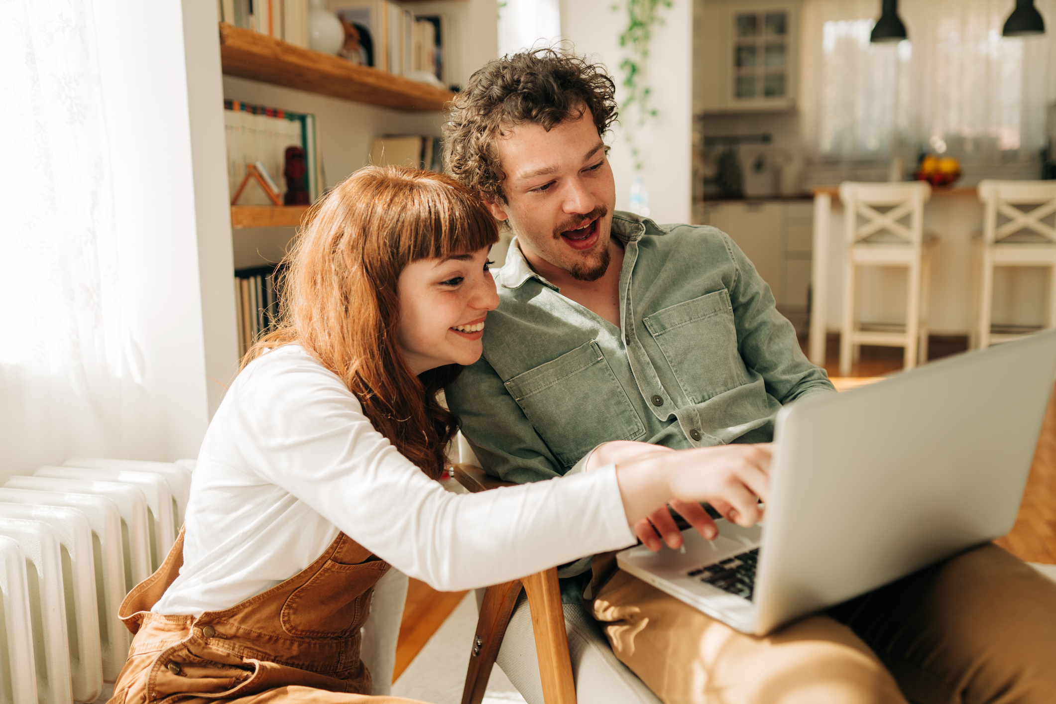 A young couple sitting closely together in a cozy home. The young woman leans in while looking at the laptop screen with interest. The young man is using the laptop as they both focus on the screen. They are browsing or discussing something online.