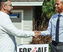2 people shaking hands in front of a for sale sign for a house