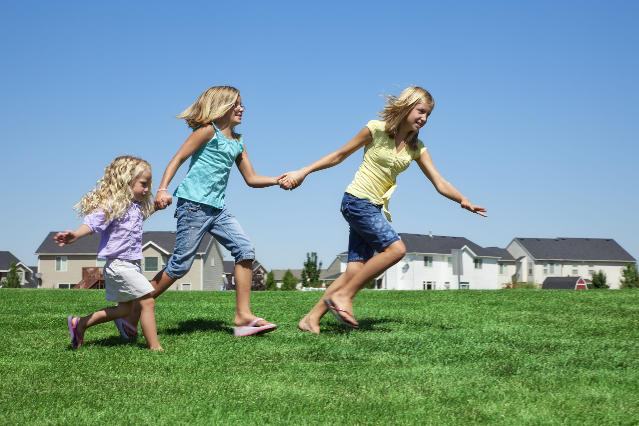 Photo of three sisters holding hands as they run through the grass of a neighborhood park.