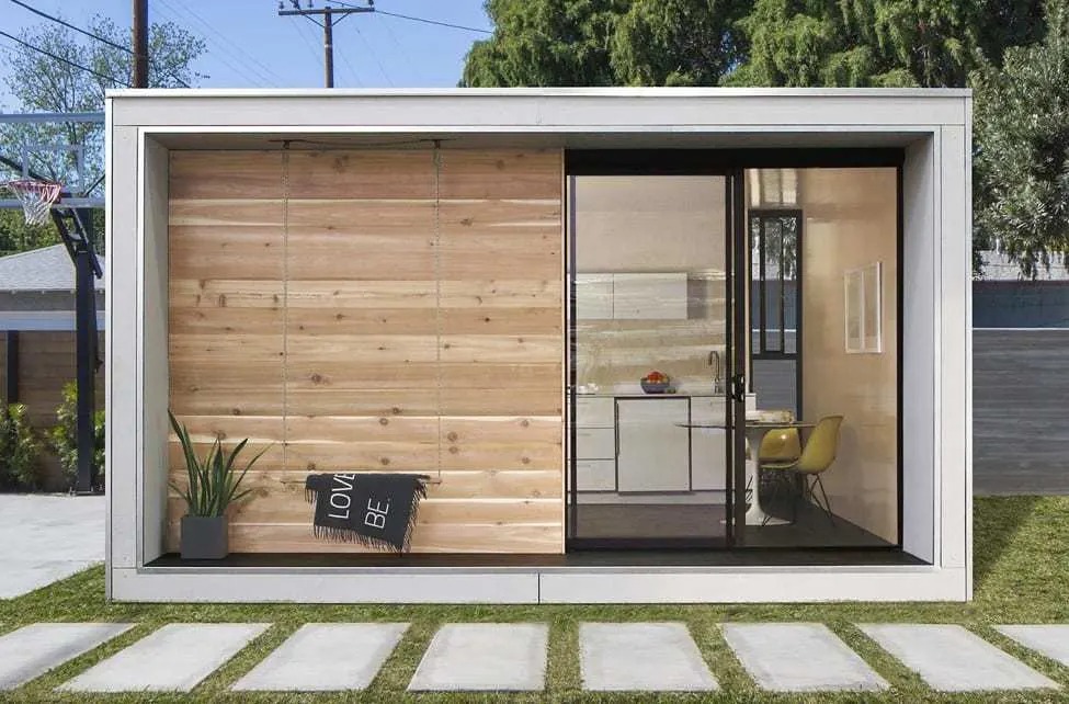 A modern, minimalist tiny house with wooden and glass walls, featuring a small kitchen and a green lawn with stone pathways.