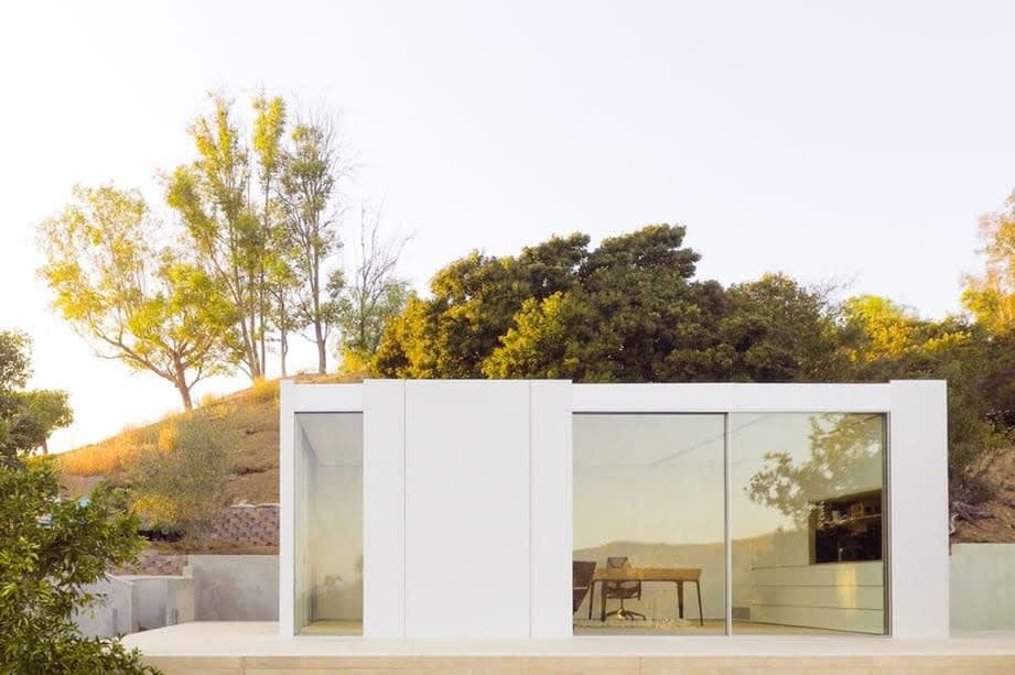 A modern, minimalist house with large glass windows, surrounded by trees and hills, reflecting a serene outdoor environment.