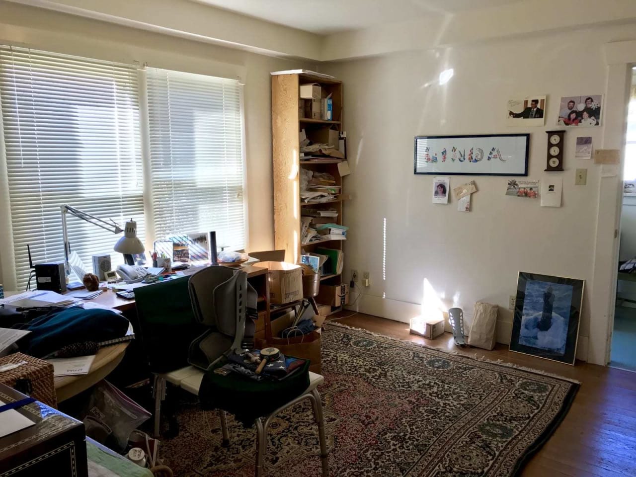 A cluttered home office with a desk, chair, and shelves filled with papers and boxes. Sunlight streams through the blinds.