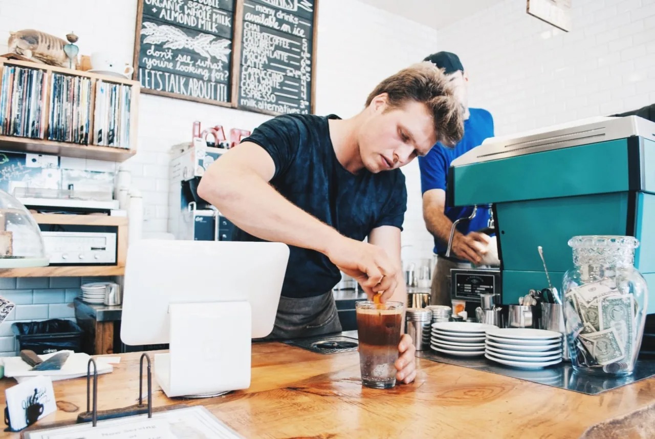 A barista prepares a drink at a coffee shop, focused on mixing ingredients in a glass. Background features a coffee machine and menu board.