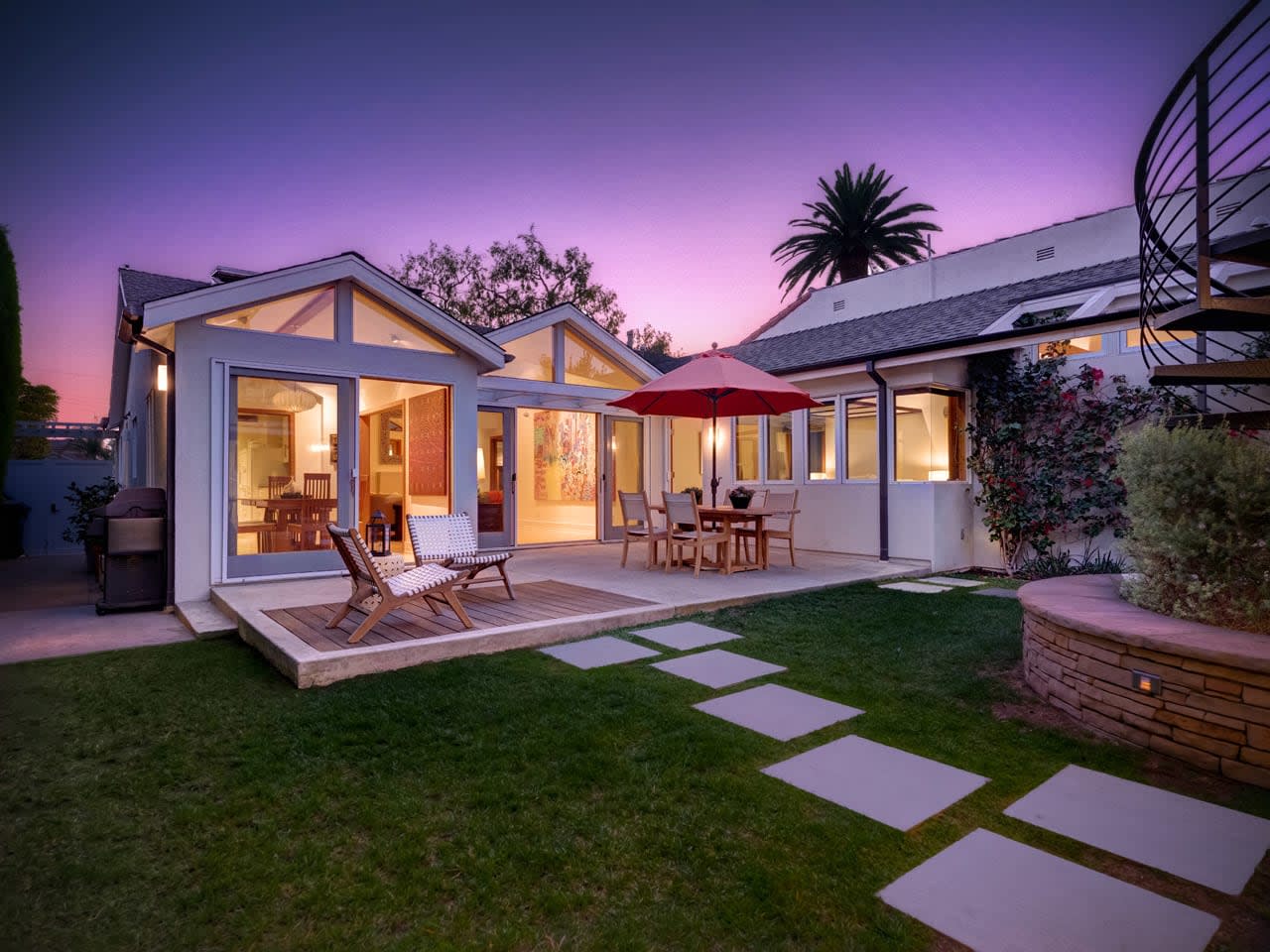 A cozy backyard with a patio, dining area under a red umbrella, and a lush lawn at sunset.