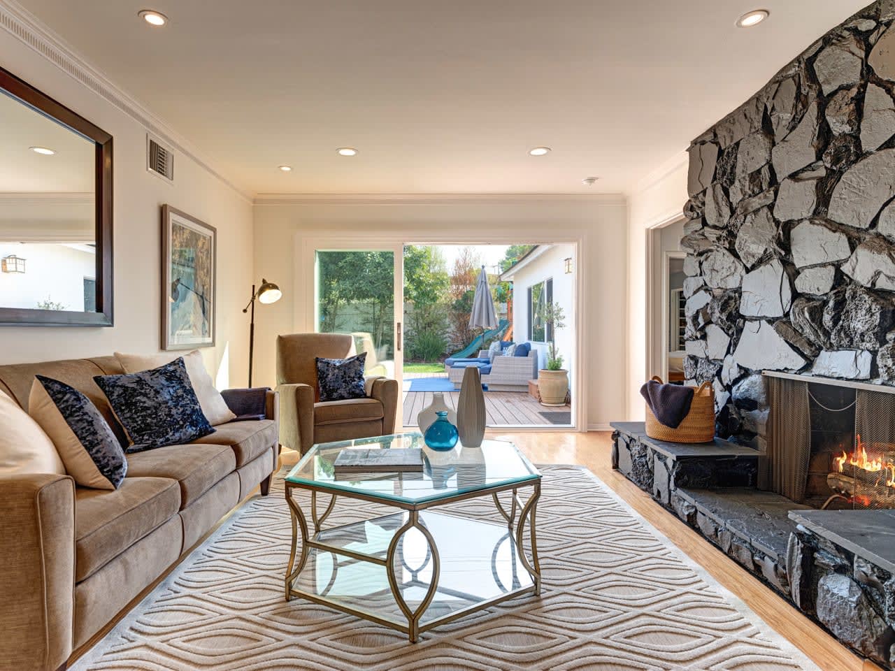 A cozy living room featuring a beige sofa, armchairs, a glass coffee table, and a stone fireplace, with large windows opening to an outdoor area.