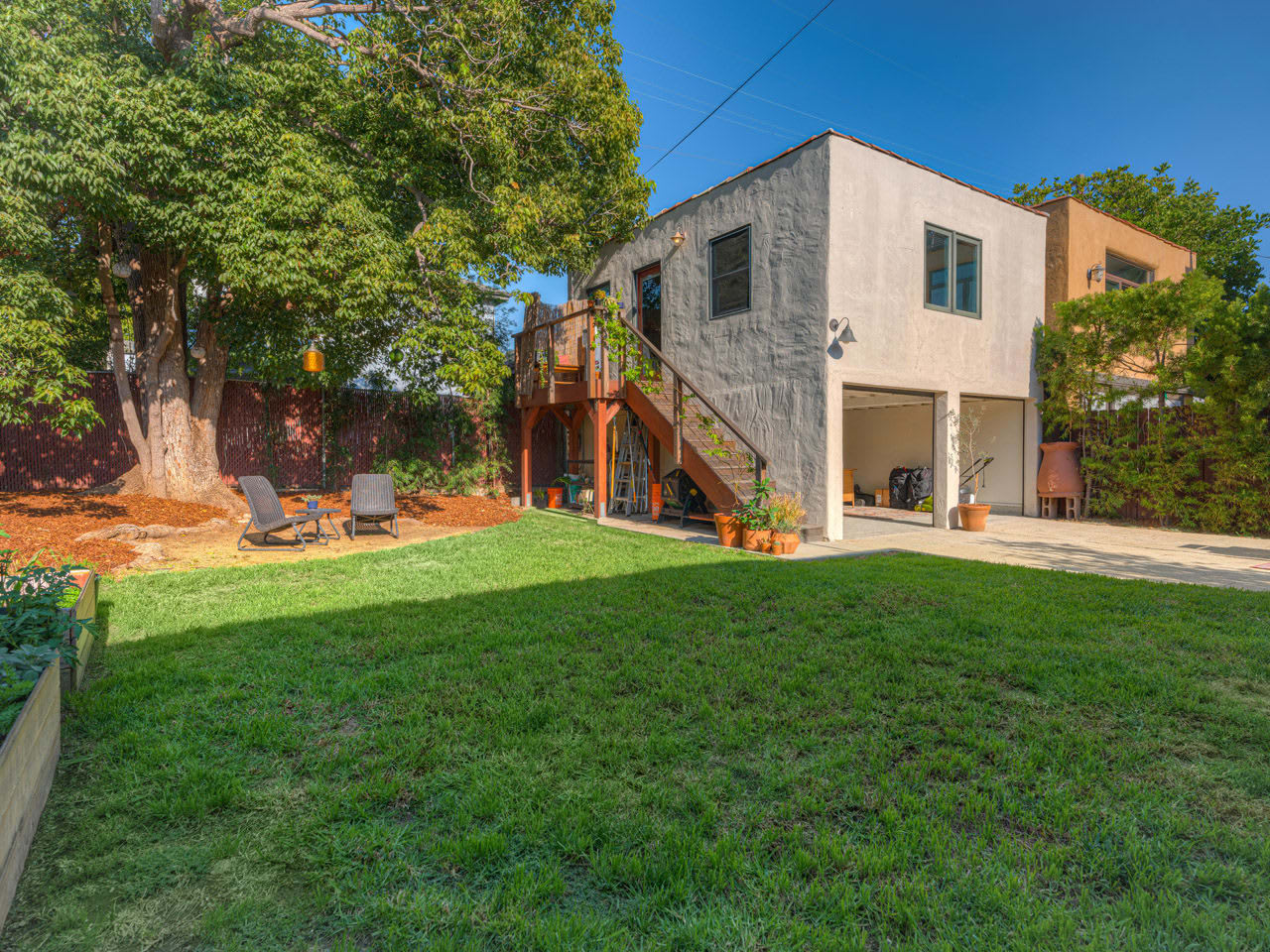 A spacious backyard featuring a green lawn, a large tree, two lounge chairs, and a two-story house with a deck.